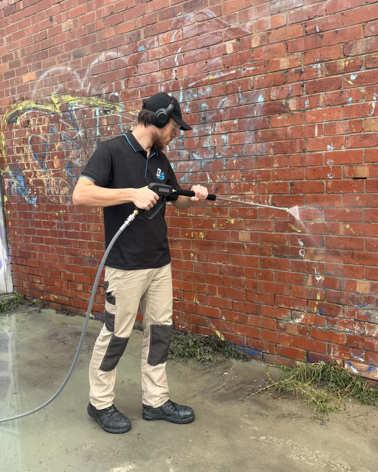 Kodi soft washing a brick wall in Graffiti Scrubbers uniform, wearing ear protection